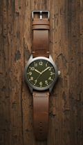 A leather-strap field watch with a robust, rugged aesthetic, positioned at the center of a weathered wood background. The...